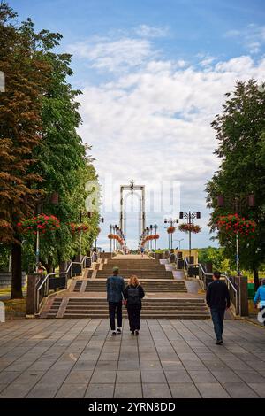 Una coppia cammina mano nella mano lungo i gradini verso un moderno arco in un parco a Kaunas, Lituania, fiancheggiato da alberi e cesti di fiori appesi sotto a. Foto Stock