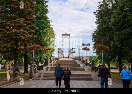 Una coppia cammina mano nella mano lungo i gradini verso un moderno arco in un parco a Kaunas, Lituania, fiancheggiato da alberi e cesti di fiori appesi sotto a. Foto Stock