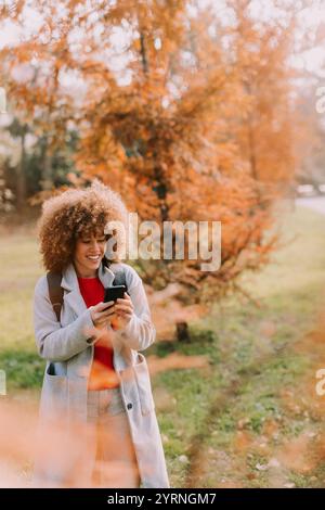 Una donna allegra si trova in un parco circondato da alberi con foglie dorate, sorridente mentre usa il telefono, immergendosi nella bellezza del giorno d'autunno. Foto Stock