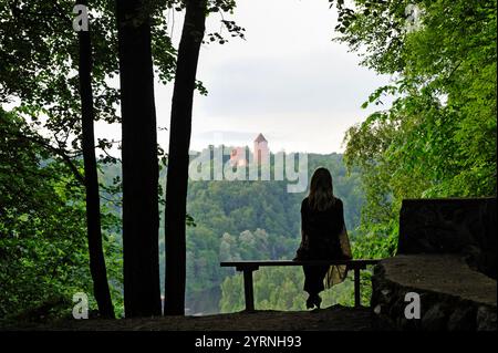 Donna seduta su una panchina che si affaccia sul fiume Gauja con sullo sfondo il castello di Turaida, intorno a Sigulda, al parco nazionale di Gauja, alla regione di Vidzeme, alla Lettonia, a Ba Foto Stock