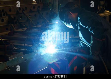 Un lavoratore esperto si concentra sulla saldatura, creando scintille e luce blu brillante. L'officina è piena di suoni di macchinari e il profumo di molte Foto Stock