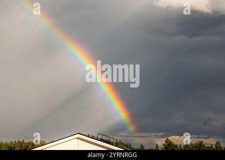 Vivace arcobaleno che si arrocca su un cielo buio e tempestoso sopra il tetto di una casa bianca con alberi circostanti in un ambiente di campagna. Svezia. Foto Stock