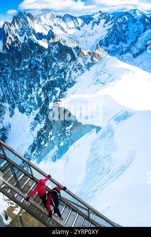 Due turisti in giacche camminano su una passerella di metallo sopra Chamonix, circondata da vaste montagne innevate. Il sole splende brillantemente sulle cime sotto a. Foto Stock