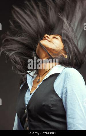 Giovane donna d'affari indiana che scuote i suoi lunghi capelli neri con gli occhi chiusi, abbraccia un momento di libertà e gioia spensierata, irradia energia e confida Foto Stock