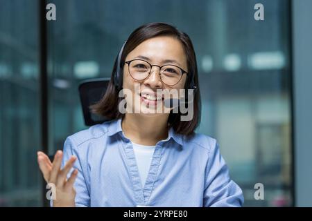 Una giovane donna asiatica allegra che indossa una cuffia lavora nell'assistenza clienti. È vestita con abbigliamento casual per affari mentre interagisce con i clienti da remoto. Foto Stock