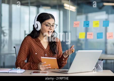 Una giovane professionista partecipa attivamente a una riunione virtuale, utilizzando il suo laptop e le cuffie per una comunicazione chiara. Tiene in mano un taccuino mentre è circondata da note adesive colorate Foto Stock