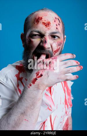Ritratto di uno zombie psicotico pazzo con il sangue sul viso che gli mordì la mano, sentendosi affamato di carne. Primo piano di una folle creatura affamata che masticava al dito, isolata sullo sfondo dello studio Foto Stock