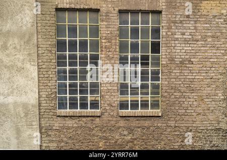 Due grandi finestre industriali con piccoli pannelli e barre metalliche sono collocate in una vecchia parete di mattoni, che mostra segni di età e usura Foto Stock