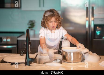 Divertenti ragazzi gemelli che aiutano in cucina con piatti di lavaggio. Bambini che si divertono con i lavori di casa. La cameriera dei bambini lava i piatti con acqua saponata. Carino Foto Stock