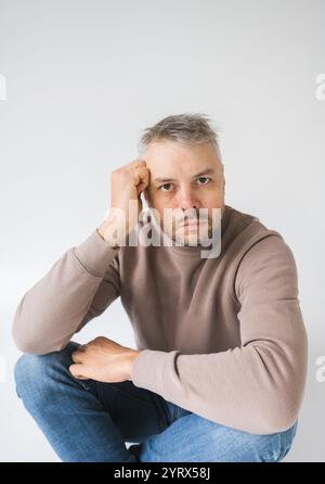 Un ritratto contemplativo di un uomo seduto, mano alla fronte, guardando direttamente la fotocamera. La sua espressione è seria e premurosa, che trasmette un Foto Stock