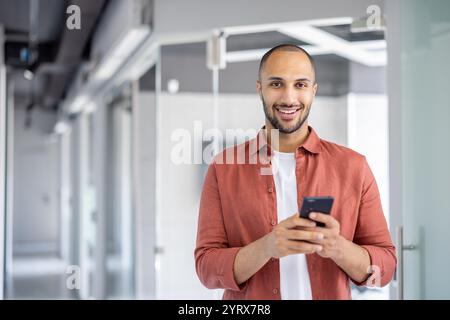 Un giovane uomo si trova in un ufficio moderno, sorridendo mentre utilizza il suo smartphone. Il suo abbigliamento casual contrasta con lo sfondo professionale, creando un'atmosfera rilassata e produttiva. Foto Stock