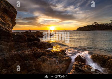 Le onde si infrangono dolcemente contro le rocce mentre il sole tramonta sulla spiaggia di Crescent Bay a Laguna Beach, gettando un caldo bagliore dorato all'orizzonte. Foto Stock