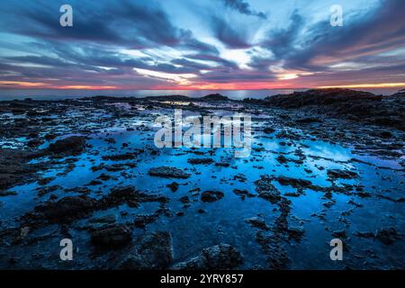 Un momento tranquillo si dispiega mentre il sole tramonta sulla spiaggia di Crescent Bay, con colori vivaci sull'oceano e sulla costa rocciosa. Foto Stock