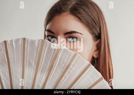 Foto in studio di una giovane donna con capelli marroni e occhi chiari che coprono parzialmente il viso con un ventilatore bianco e beige, creando un senso di mistero A. Foto Stock