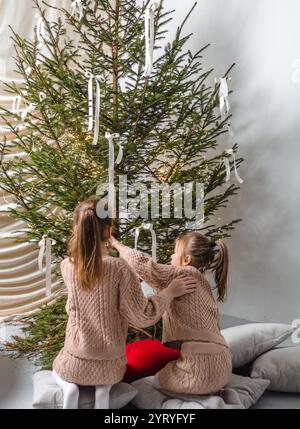 Due ragazze con abiti in maglia a cavo abbinati decorano un albero di Natale con nastri bianchi. Foto Stock