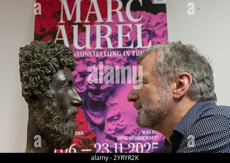 Treviri, Germania. 4 dicembre 2024. Marcus Reuter, direttore del Rheinisches Landesmuseum di Treviri, guarda nella macchina fotografica di fronte a un poster per la mostra di stato accanto a una replica del capo dell'imperatore romano Marco Aurelio. La mostra al Rheinisches Landesmuseum e allo Stadtmuseum Trier si svolge dal 15 giugno al 23 novembre 2025. Credito: Harald Tittel/dpa/Alamy Live News Foto Stock