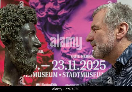 Treviri, Germania. 4 dicembre 2024. Marcus Reuter, direttore del Rheinisches Landesmuseum di Treviri, guarda nella macchina fotografica di fronte a un poster per la mostra di stato accanto a una replica del capo dell'imperatore romano Marco Aurelio. La mostra al Rheinisches Landesmuseum e allo Stadtmuseum Trier si svolge dal 15 giugno al 23 novembre 2025. Credito: Harald Tittel/dpa/Alamy Live News Foto Stock