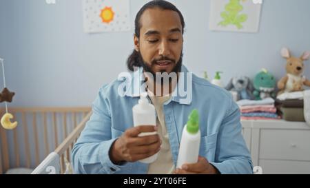 Uomo con barba che tiene in mano prodotti per bambini in un'accogliente camera da letto, che mostra un'atmosfera nutriente piena di giocattoli, pareti pastello e una culla in legno. Foto Stock