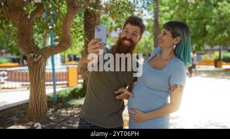 Uomo e donna che si godono una videoconferenza in un parco urbano, puntando alla sua pancia incinta, festeggiando insieme la gravidanza all'aperto in una giornata di sole Foto Stock