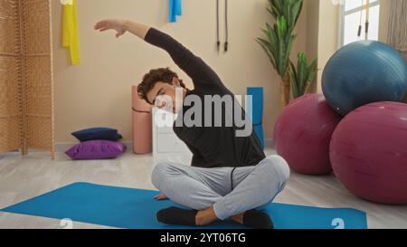 Giovane uomo che si allunga sul tappetino in palestra con palle da ginnastica in un luminoso ambiente interno, che mette in mostra fitness e flessibilità. Foto Stock