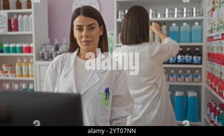 Due farmaciste che lavorano insieme in un negozio di farmacia vestite di camici bianchi da laboratorio, una guarda la fotocamera mentre l'altra prepara il prodotto Foto Stock