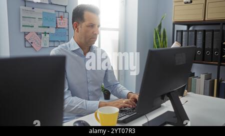 Uomo ispanico che lavora diligentemente su un computer in un ufficio organizzato con note strategiche, leganti e una tazza di caffè gialla sulla scrivania. Foto Stock