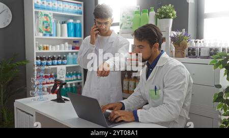 Farmacisti che lavorano insieme in un computer in un negozio di farmacia con scaffali di prodotti sanitari dietro di loro, uno che parla al telefono mentre l'altro t Foto Stock