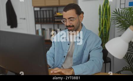 Bellissimo giovane ispanico con baffi che lavora al chiuso in ufficio con un computer, concentrato e professionale Foto Stock