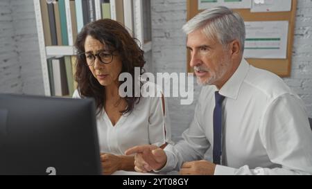 Uomo e donna che collaborano a un computer in un ufficio affollato, circondati da documenti, concentrandosi sul lavoro di squadra, sulla comunicazione e sulla risoluzione dei problemi Foto Stock