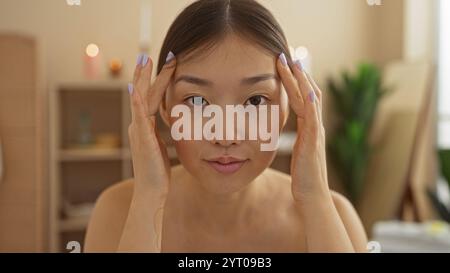 Giovane, bella, asiatica, donna in un centro benessere, che guarda direttamente la macchina fotografica toccando il suo viso in un ambiente tranquillo e al coperto con morbidezza Foto Stock