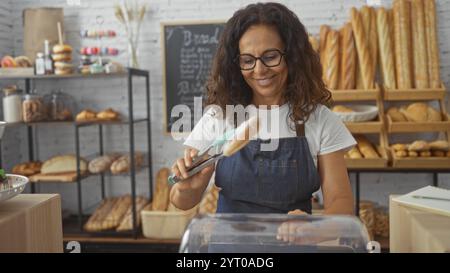 Donna che lavora in una panetteria, indossa un grembiule e pinze, mette in mostra il pane, sorride tra scaffali pieni di vari prodotti da forno Foto Stock