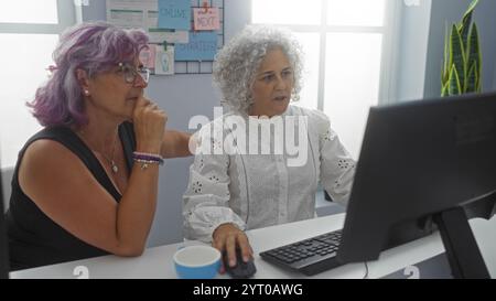 Donne di mezza età che collaborano in un ufficio, intensamente focalizzate sullo schermo di un computer, mostrano una collaborazione lavorativa in un moderno lavoro d'interni Foto Stock