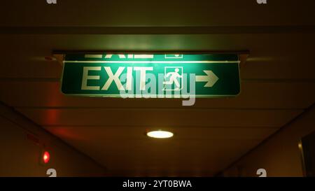 Il segnale di uscita illuminato che si illumina all'interno mostra la direzione in un corridoio poco illuminato, evidenziando il design di sicurezza e la consapevolezza delle emergenze con il suo verde brillante Foto Stock
