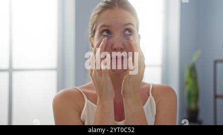 Giovane donna bionda che applica crema per la cura della pelle all'interno di una sala del centro benessere spa con luce naturale soffusa dalle finestre Foto Stock