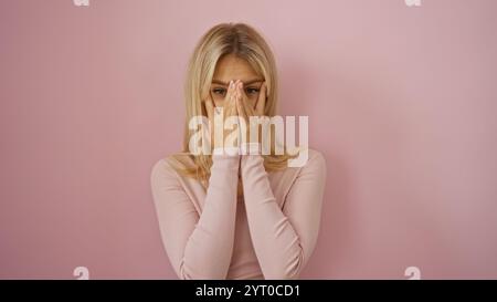 Donna che ricopre il viso con le mani davanti allo sfondo rosa trasuda bellezza e fascino giovanile in un ambiente isolato. Foto Stock