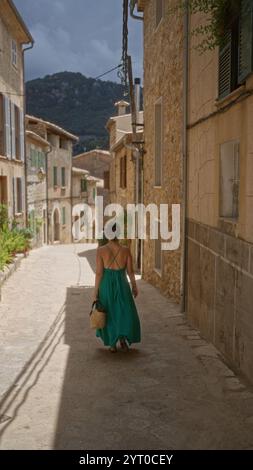 Donna che cammina per le strette strade di valldemossa a maiorca, spagna, indossando un abito verde con montagne sullo sfondo Foto Stock