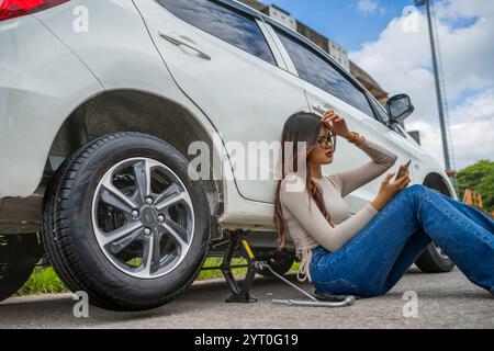 Una donna sta utilizzando il suo smartphone mentre cambia una ruota sgonfia sul suo SUV Foto Stock