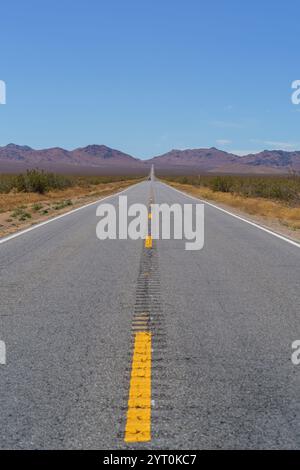 Una lunga e dritta strada asfaltata con linee gialle si estende verso montagne lontane e basse sotto un cielo azzurro nel deserto. Foto Stock