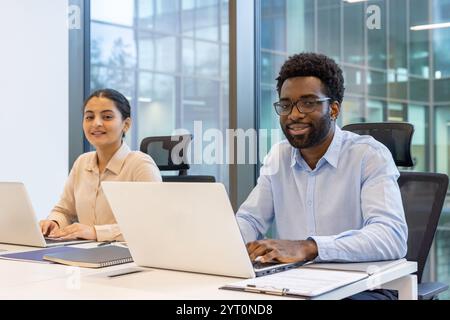 Un giovane uomo e una donna sono seduti a una scrivania che lavora su un computer portatile in un ufficio moderno. Sembrano concentrati e impegnati, rappresentando il lavoro di squadra e la produttività in un ambiente professionale. Foto Stock