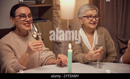 Due donne vestite con comodi maglioni caldi, sedute a tavola in un soggiorno illuminato, bevendo vino bianco, parlando, sorridendo Foto Stock