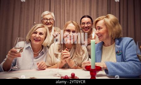 Gruppo di cinque anziane donne sorridenti, che si riuniscono intorno a un tavolo decorato con candele in serata accogliente, ridendo, parlando e bevendo vino Foto Stock