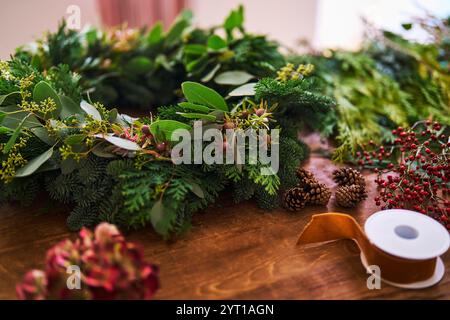 Primo piano di Foliage con Fiori, bacche e cono di pino utilizzati per decorare la corona natalizia sul tavolo a casa con From Ribbon Foto Stock
