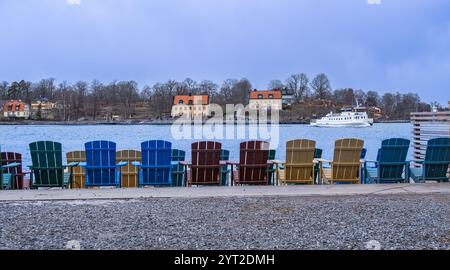 Stoccolma, Svezia - 5 marzo 2023: Colorate sedie Adirondack di fronte a un tranquillo lago con case e una barca sullo sfondo. Foto Stock