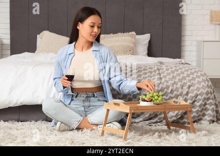 Giovane donna asiatica seduta sul pavimento con un bicchiere di vino e mangiando uva dal vassoio a casa Foto Stock