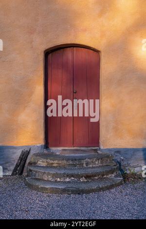 Una porta in legno rosso rustico con una parte superiore ad arco, adagiata in una parete di gesso giallo, circondata da gradini in pietra Foto Stock