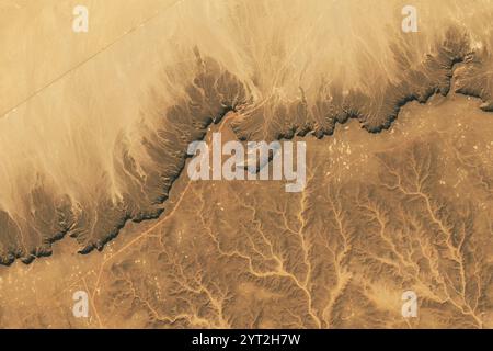 Libia. 17 novembre 2024. Torreggiante sul deserto nella Libia sud-occidentale, l'altopiano di Messak Settafet assomiglia al muro di una gigantesca fortezza, con la sua pietra arenaria scura e resistente all'erosione che separa il Mare di sabbia di Ubari a nord e il Mare di sabbia di Marzuq a sud. Gli oli-2 su Landsat 9 catturarono queste immagini del plateau. Il grande affioramento di roccia del Cretaceo presenta una acuta scarpata sul lato nord. La scogliera, a circa 1.000 metri di altezza, è allineata con una sottile ombra scura nell'immagine in alto. I detriti scuri delle cascate sono visibili a nord. Si noti che un'illusione ottica calle Foto Stock