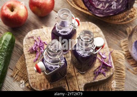 Tre bottiglie di succo di cavolo viola con mele fresche e cetriolo su un tavolo Foto Stock