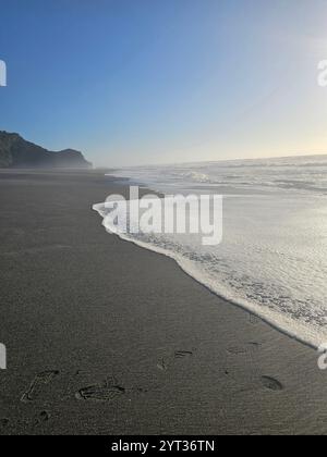 Tranquillo paesaggio costiero con dolci onde che scivolano sulla spiaggia sabbiosa, illuminato dalla luce soffusa del sole sotto un cielo azzurro. Foto Stock