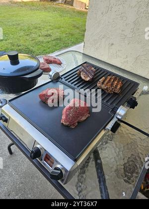 Grigliate all'aperto con bistecche cotte e crude su un piatto, circondate da un cortile erboso e da un patio. Cattura un momento di barbecue informale. Foto Stock