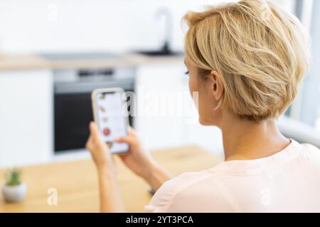 Donna caucasica che usa lo smartphone per ordinare il cibo online in un ambiente moderno. Donna bionda con capelli corti che sfogliano il sito Web di consegna. L'immagine riflette la comodità, la tecnologia e lo stile di vita digitale. Foto Stock